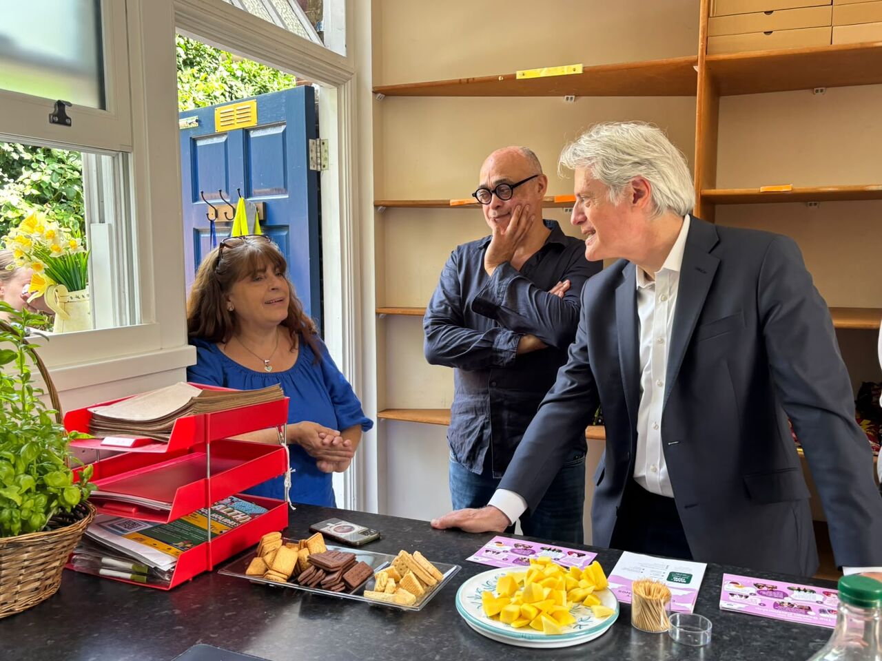 This image shows three people gathered around a dark counter or reception area indoors, appearing to be in an informal, friendly conversation. People Present Left: A woman with long brown hair, wearing a royal blue top, standing near an open blue door. She has her hands together in front of her and is looking towards the man on the right. Middle: A man with a bald head, dark-rimmed glasses, and a dark long-sleeved shirt leaning back against wooden shelving units, holding his hand to his cheek while listening. Right: A man with grey hair, wearing a navy blue suit jacket over a open-collared white shirt, leaning forward over the counter with his hand resting on the surface.