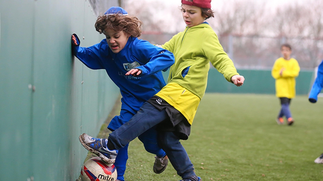 Two young players are locked in a close-contact struggle for the ball along the perimeter wall of the pitch. One player in a bright blue long-sleeved jersey has their foot on the ball, shielding it from an opponent in a lime-green hoodie and yellow shorts.