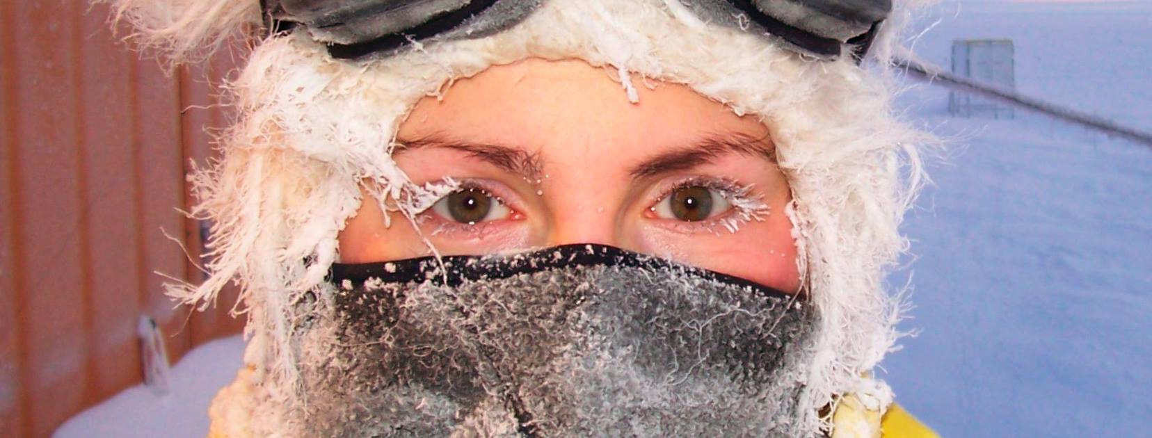 A close-up of a person's face, wrapped up against extreme cold. Only the eyes and eyebrows are visible between a fur-trimmed hood and a dark face covering, both crusted with frost and ice. Even the eyelashes and stray hairs are frozen white. Snow and a hint of a reddish building fill the blurred background under cold, pale light.