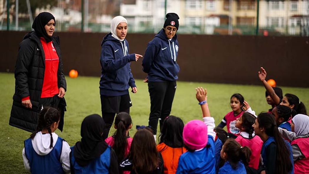 Three female coaches are leading a session with a group of young girls on an outdoor artificial turf pitch. One coach, wearing a white headscarf and a navy hoodie, is actively gesturing and speaking to the attentive player
