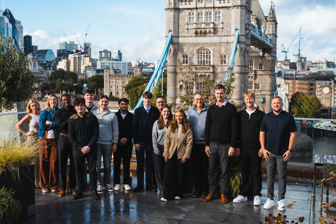 People & Composition: A group of 15 people are standing together in a line outdoors on an open-air terrace, smiling at the camera. They are dressed in casual to smart-casual autumn attire, including jackets, sweaters, collared shirts, and trousers. Iconic Landmarks: The photo is taken against the backdrop of London's Tower Bridge, with its distinctive stone towers and blue suspension cables rising directly behind the group. To the left in the mid-ground, the historic Tower of London is visible. City Skyline: Further in the background on the left, the modern skyline of the City of London can be seen, including recognizable skyscrapers like The Gherkin (30 St Mary Axe). Setting & Environment: The group is on a wet, tiled rooftop terrace with potted greenery and small trees framing the sides. The weather appears bright with partial sun and blue skies overhead.