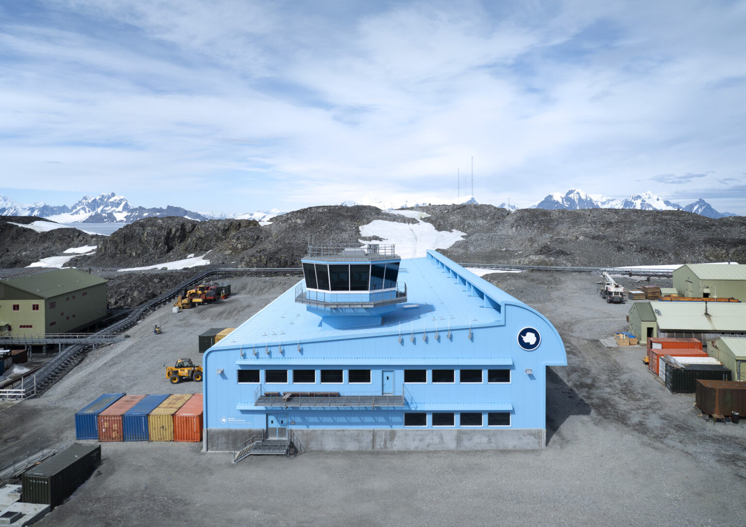 The image shows the Discovery Building at the Rothera Research Station in Antarctica. It is a large, two-storey modern facility characterised by its striking bright blue exterior, designed to stand out against the stark, monochromatic polar landscape. Architectural Details Structure: The building has a long, aerodynamic shape with a distinct curved roofline on one side, which serves as a wind deflector to prevent snow build-up. Control Tower: A multi-sided, glass-walled control tower sits atop the flat section of the roof, providing a 360-degree view of the station and the surrounding runway and wharf areas. Windows: Rows of rectangular windows line the upper and lower floors, designed to maximise natural light while maintaining high thermal efficiency. Foundation: The building is set on a solid concrete plinth, elevating the metal-clad structure slightly above the rocky, permafrost ground. Surroundings and Context Landscape: The building is situated in a rugged, rocky environment with sparse patches of snow. In the background, jagged, snow-capped mountains and glaciers rise under a pale blue, slightly cloudy sky. Logistics: To the left and right of the building, several colourful shipping containers (blue, orange, and red) and heavy machinery, including a yellow telehandler, indicate ongoing operational and construction activity. Branding: The British Antarctic Survey (BAS) logo—a circular emblem featuring a map of Antarctica—is prominently displayed on the side of the building. The overall impression is one of cutting-edge engineering and resilience, a high-tech "hub" sitting in one of the most remote and extreme environments on Earth.