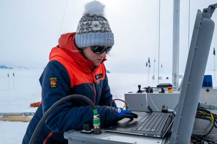 Emma Fisher works intently at a laptop propped on a grey control box, wearing a heavy navy and orange jacket, a knitted bobble hat and dark sunglasses. Thick cables with heavy-duty connectors run into the equipment, and a larger control unit with joysticks and a red emergency stop button stands to the right. Behind her stretches a flat expanse of Antarctic snow and ice under a pale, overcast sky, with marker flags receding into the distance.