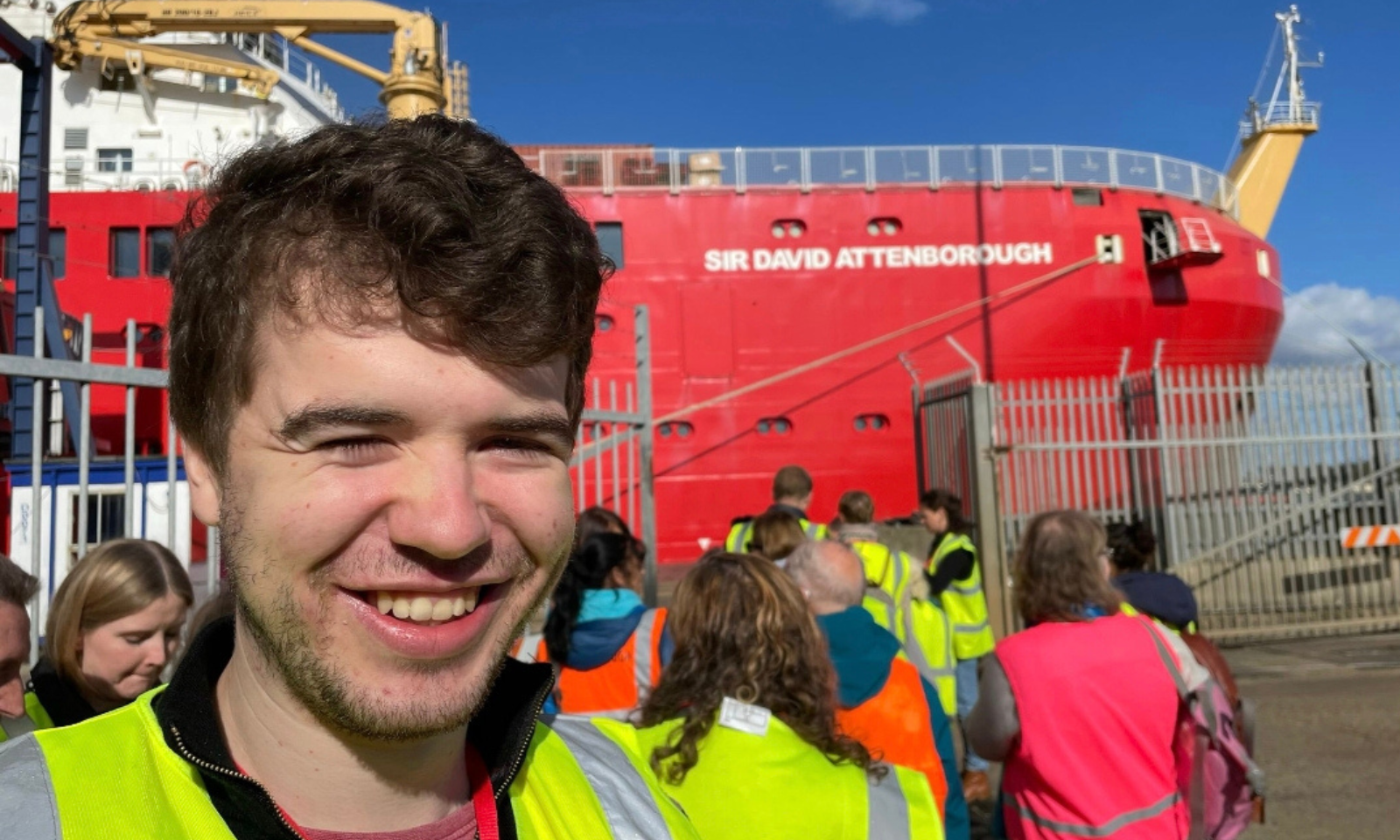 This is a bright, outdoor photograph capturing a smiling young man in the foreground with a large, distinctive research vessel docked in the background.  Foreground The Subject: A young man with brown, wavy hair and a light beard/stubble is smiling broadly at the camera. He is wearing a high-visibility yellow safety vest over a dark zip-up collar and a reddish undershirt.  Other People: To his left (the viewer's bottom-left), the top of a woman&rsquo;s head with blonde hair is visible, looking downwards.  Midground A Group of Visitors: A small crowd of people is gathered behind a metal security fence. Many of them are facing away from the camera towards the ship. Several individuals in this group are wearing high-visibility gear, including yellow safety vests, orange vests, and one person in a bright pink vest.  Security Gate: A metal palisade fence separates the foreground area from the dock where the ship is moored.  Background The Vessel: Dominated by a massive, bright red ship. Printed clearly on the side of the hull in white, capital block letters is the name "SIR DAVID ATTENBOROUGH" (the famous British polar research ship).  Ship details: A yellow crane structure is visible on the upper deck to the left, and a yellow mast is visible on the bow to the right.  The Sky: The weather appears clear and sunny with a bright blue sky and a few light, wispy clouds.