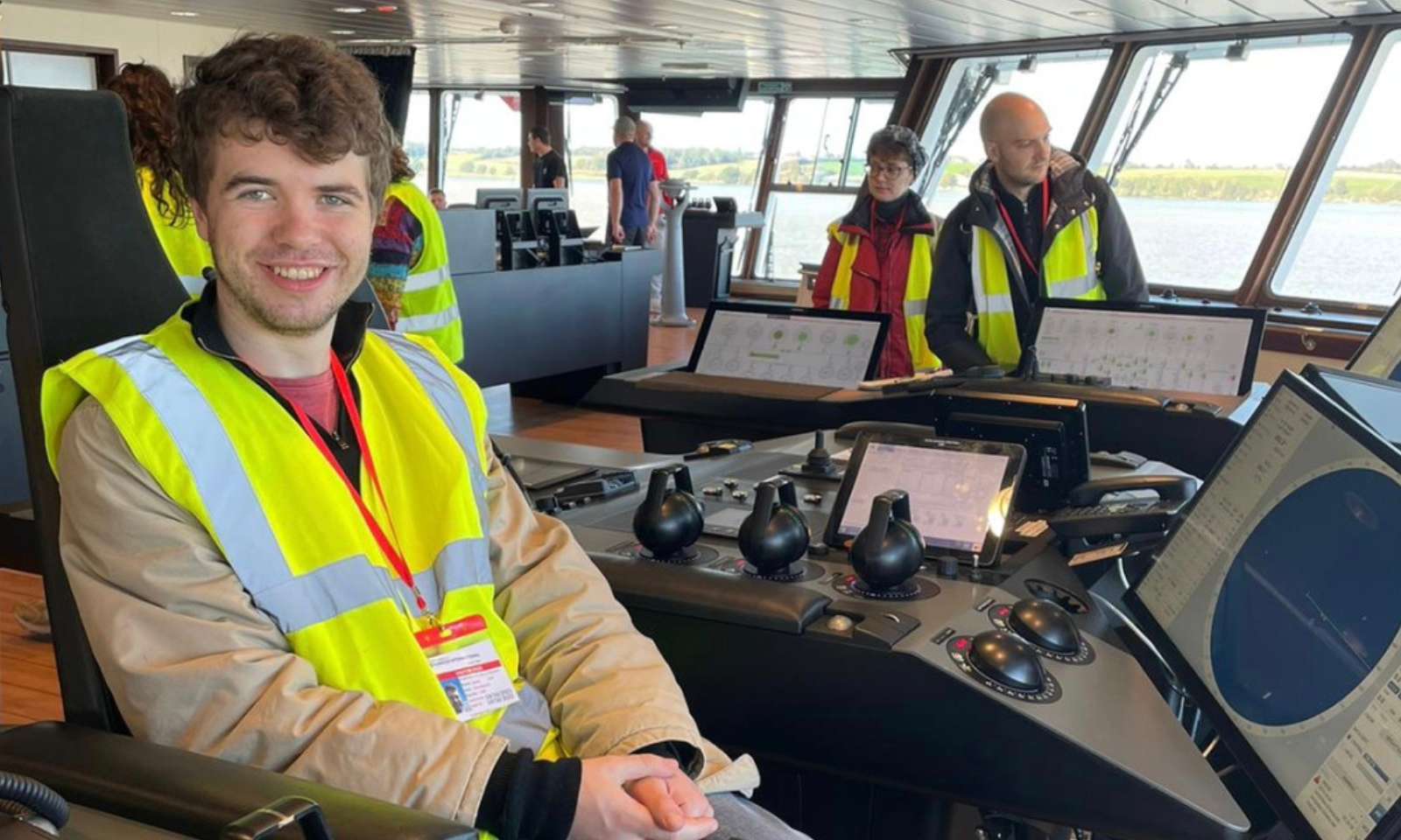 This is an indoor photograph capturing the same young man, this time seated inside the bridge of a vessel (likely the Sir David Attenborough from the previous photo), surrounded by navigation equipment and other visitors.  Following your preference, this description uses UK spelling.  Foreground The Subject: The young man with brown, wavy hair is sitting in a large, dark captain's or officer's chair on the left side of the frame. He is smiling at the camera with his hands clasped in his lap.  Clothing and Gear: He is wearing a beige jacket under a high-visibility yellow safety vest with reflective strips. A red lanyard hangs around his neck, holding a visitor or identification badge.  Control Console: In front of him is a large, modern black console equipped with multiple control levers (thruster/propulsion grips), buttons, and dials. A digital tablet-style screen is mounted in the centre, and a larger angled navigation display showing a radar or map interface is visible on the right.  Midground and Background Other Visitors: Several other people are visible on the spacious bridge, also wearing high-visibility yellow vests, indicating a guided tour.  To the immediate right, a bald man and a woman with glasses are standing near another set of control consoles, looking downwards or towards the equipment.  Further back, more individuals are standing near the central control stations.  The Bridge Interior: The room features wood-effect flooring, sleek black console units, and a row of large, forward-slanted windows that line the front of the bridge.  The View Outside: Through the windows, calm water is visible, with a green coastline or riverbank stretching across the horizon under a bright sky.