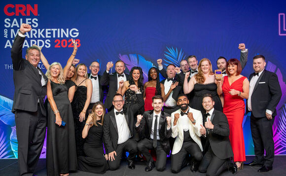 Event & Branding: A large group is posing in front of a blue backdrop featuring tropical leaf graphics and white-and-red text reading "CRN SALES AND MARKETING AWARDS 2023". People & Pose: The photo features a lively group celebrating on stage, with many raising their fists, cheering, giving thumbs-up, or holding champagne glasses. One woman near the center-right is holding up an award trophy. Attire: Everyone is dressed in formal evening wear (black-tie event style). The men are wearing tuxedos or suits with bowties or ties—including one man in a cream/white dinner jacket—and the women are wearing evening gowns and cocktail dresses in black, red, and other dark shades.