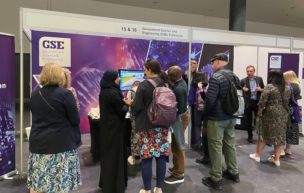 Several people are gathered around the stand, engaging with exhibitors. On the left, a woman in a black hijab is speaking with visitors, while on the right, a man in a suit is gesturing while talking to a small group.