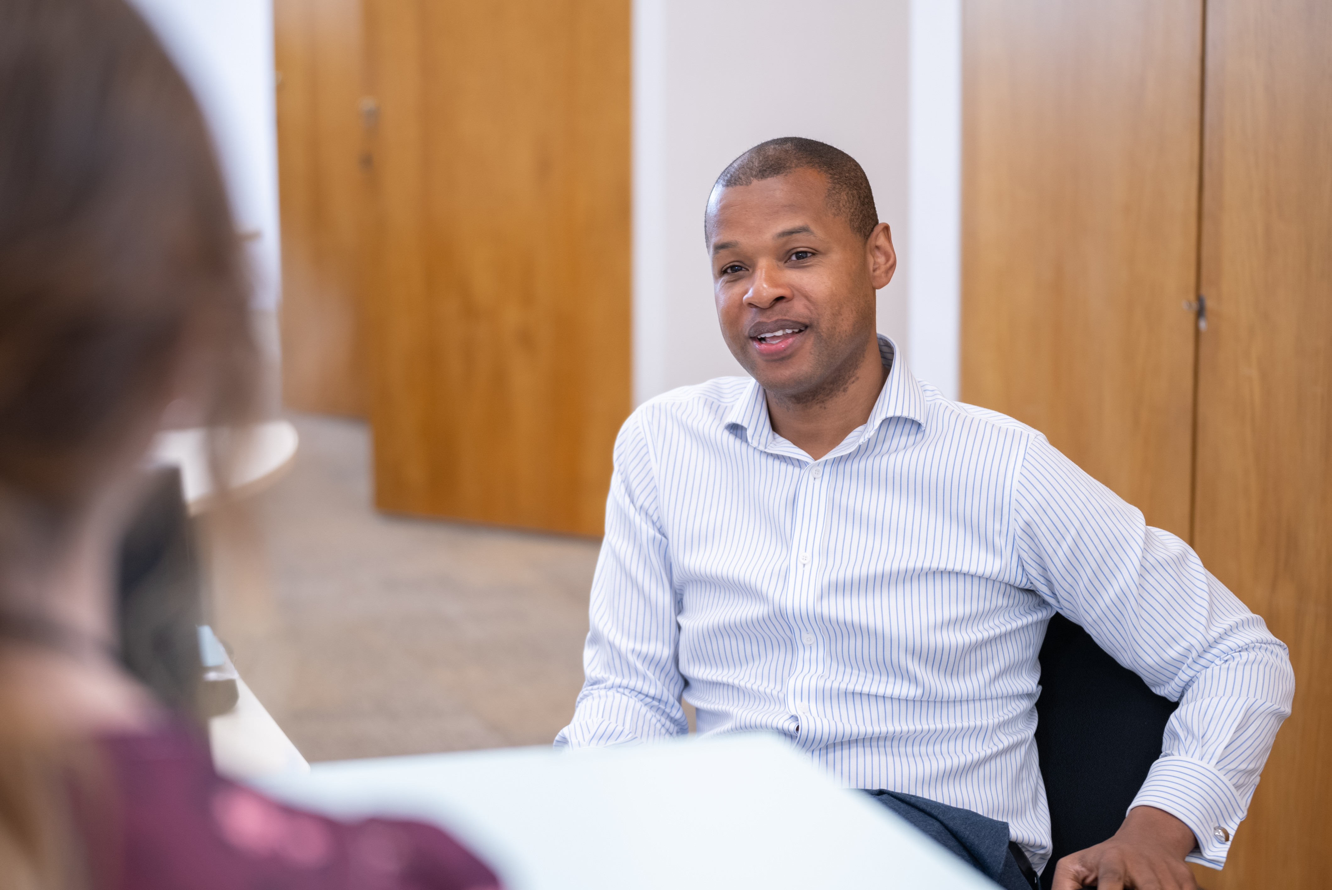 This image depicts a professional workplace conversation in an office setting: People & Interaction: On the right, a Black man with short hair is seated and leaning back comfortably in an office chair. He is wearing a white button-down shirt with light blue vertical stripes and appears to be speaking with an open, pleasant expression. In the foreground on the left, the back of another person's head and shoulder is visible, wearing a dark purple patterned garment, suggesting a two-person meeting or discussion across a desk. Setting & Details: The background features modern office decor with light wooden doors, neutral-toned walls, and soft lighting. The overall atmosphere feels approachable, collaborative, and professional.