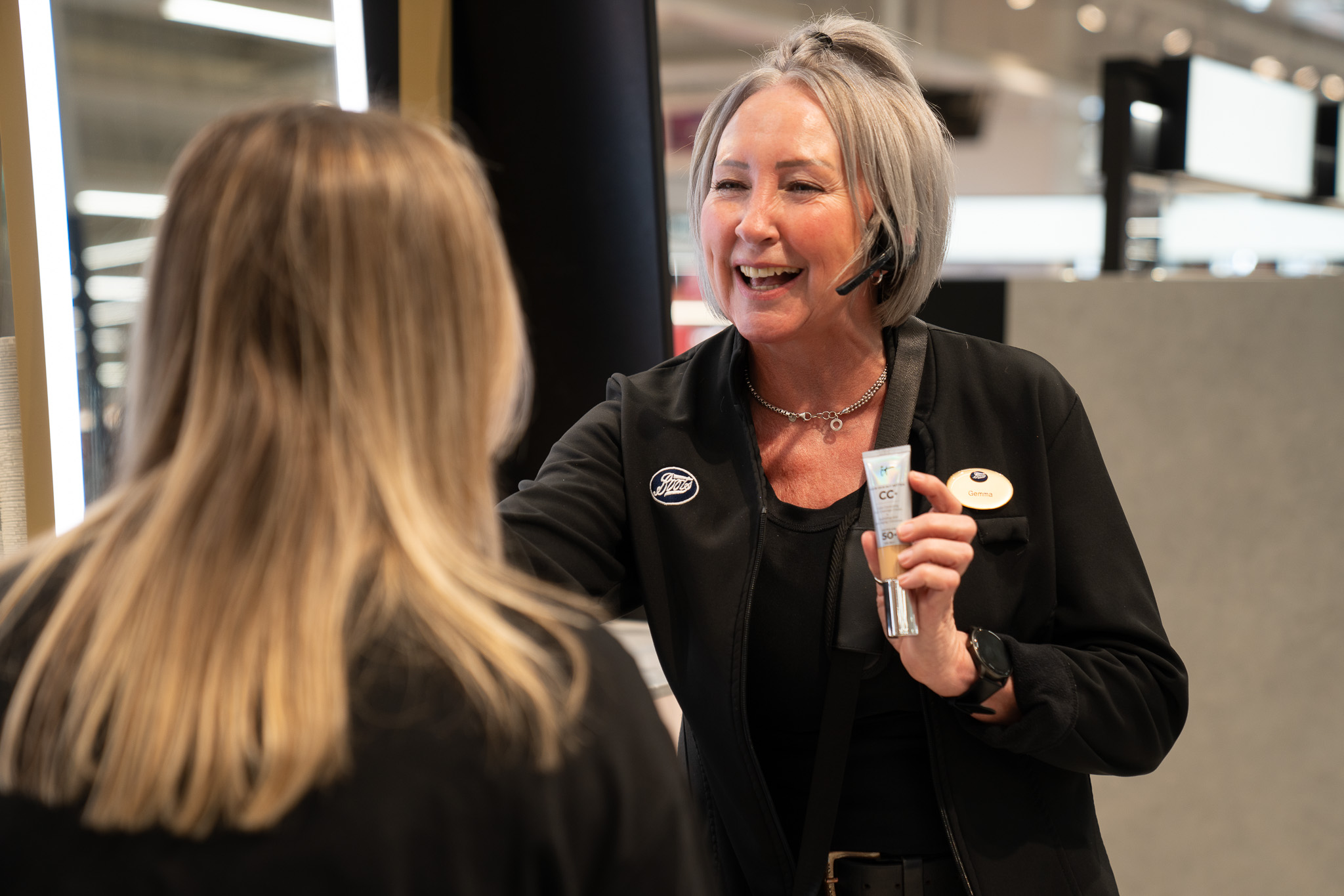 This image shows a customer interaction inside a brightly lit retail store, likely a beauty counter or pharmacy section:  People & Interaction:  On the right, a smiling woman with short silver-grey hair wears a black jacket featuring a Boots logo badge on the chest and an oval name tag. She is wearing a headset and holding up a tube of cosmetic product (a CC+ cream) to present to a customer.  On the left, viewed from behind, is a customer with straight blonde hair, listening attentively as the staff member demonstrates the product.  Setting & Details:  The environment appears to be a modern retail space, with bright vanity lighting on a mirror visible behind the customer's shoulder and softly blurred store aisles and displays in the background.  The store staff member wears a necklace, a black wristwatch, and a black strap slung across her chest.