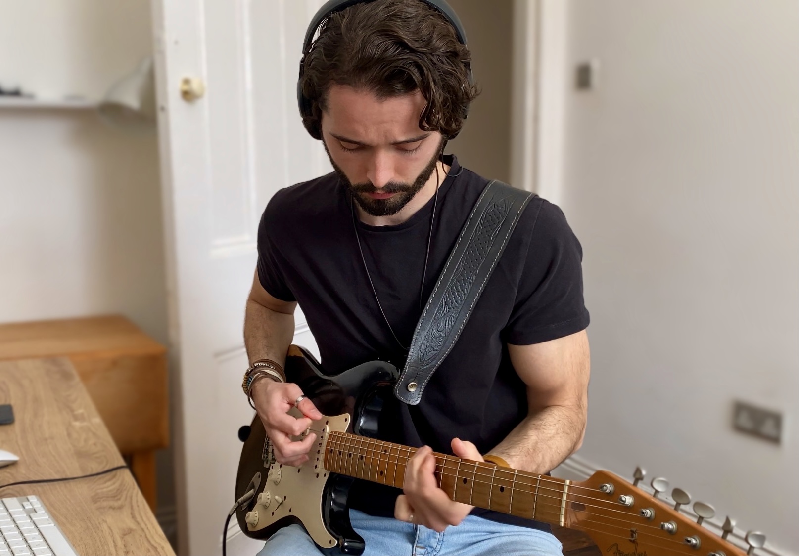 Subject: David Smith, a man with dark, wavy hair and a beard, is captured mid-performance playing an electric guitar. Attire & Accessories: He is wearing a plain black crew-neck t-shirt, light blue jeans, over-ear headphones, and beaded bracelets on his left wrist. A dark leather guitar strap with an embossed pattern crosses his left shoulder. Instrument: He is holding a black electric guitar with a white pickguard and a light-coloured wooden fretboard. His fingers are positioned on the frets and strings as if playing chords. Setting: He is indoors, seated near a wooden desk where part of a computer keyboard is visible. The background shows a neutral-toned room with a white door and a wall socket.