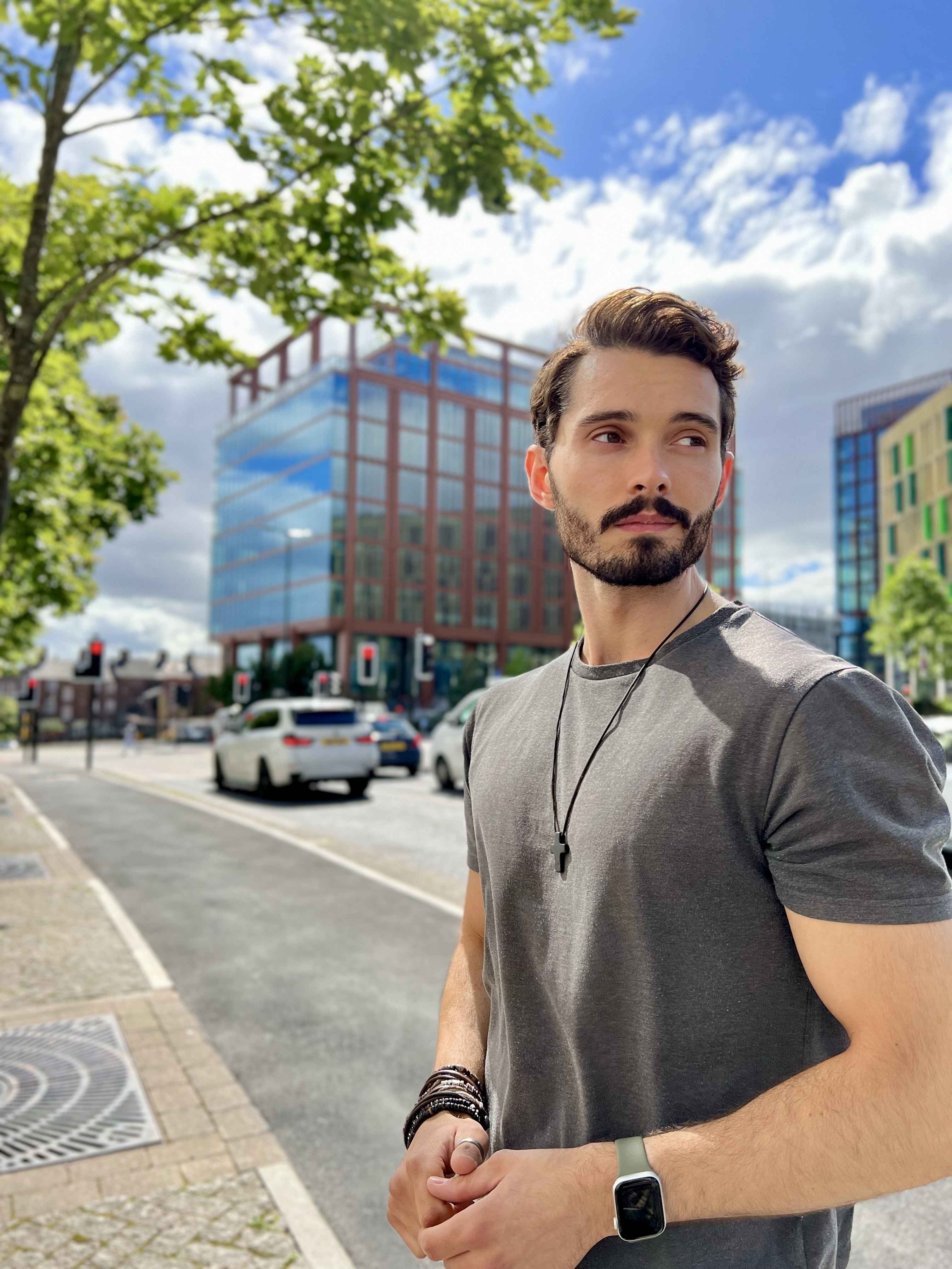 Subject: David is standing outdoors on a city pavement, turned slightly in profile and looking off to his left. He has short brown hair styled upward and a full beard and moustache. Attire & Accessories: He is wearing a grey t-shirt and a dark pendant necklace with a cross. On his wrists, he wears stacked dark leather or beaded bracelets, a silver ring, and a light-green smartwatch or fitness tracker. Background & Setting: The image features an urban street scene under a partly cloudy blue sky. In the background are modern city buildings, streetlights, traffic, and leafy green trees.