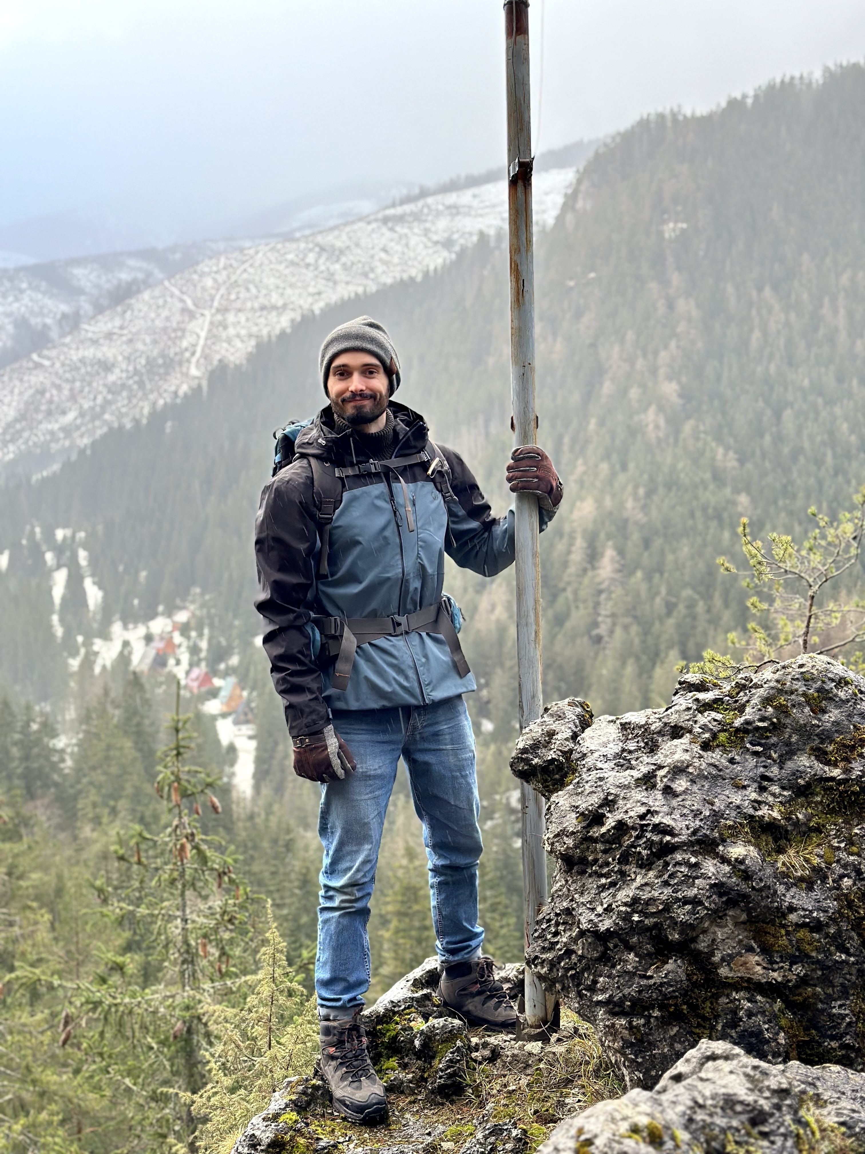 Subject: David is standing outdoors on a rocky vantage point in a mountainous landscape, smiling at the camera while holding onto a tall metal pole with his left hand. Attire & Gear: A grey knitted beanie hat. A two-tone blue and black hooded outdoor jacket worn over a dark roll-neck top. A hiking backpack secured with chest and waist straps. Light-blue jeans, brown gloves, and sturdy dark hiking boots. Background & Setting: He is surrounded by a rugged, scenic mountain landscape featuring dense pine forests, misty valleys, and patches of snow on the distant slopes. A small cluster of cabins is visible down in the valley below.
