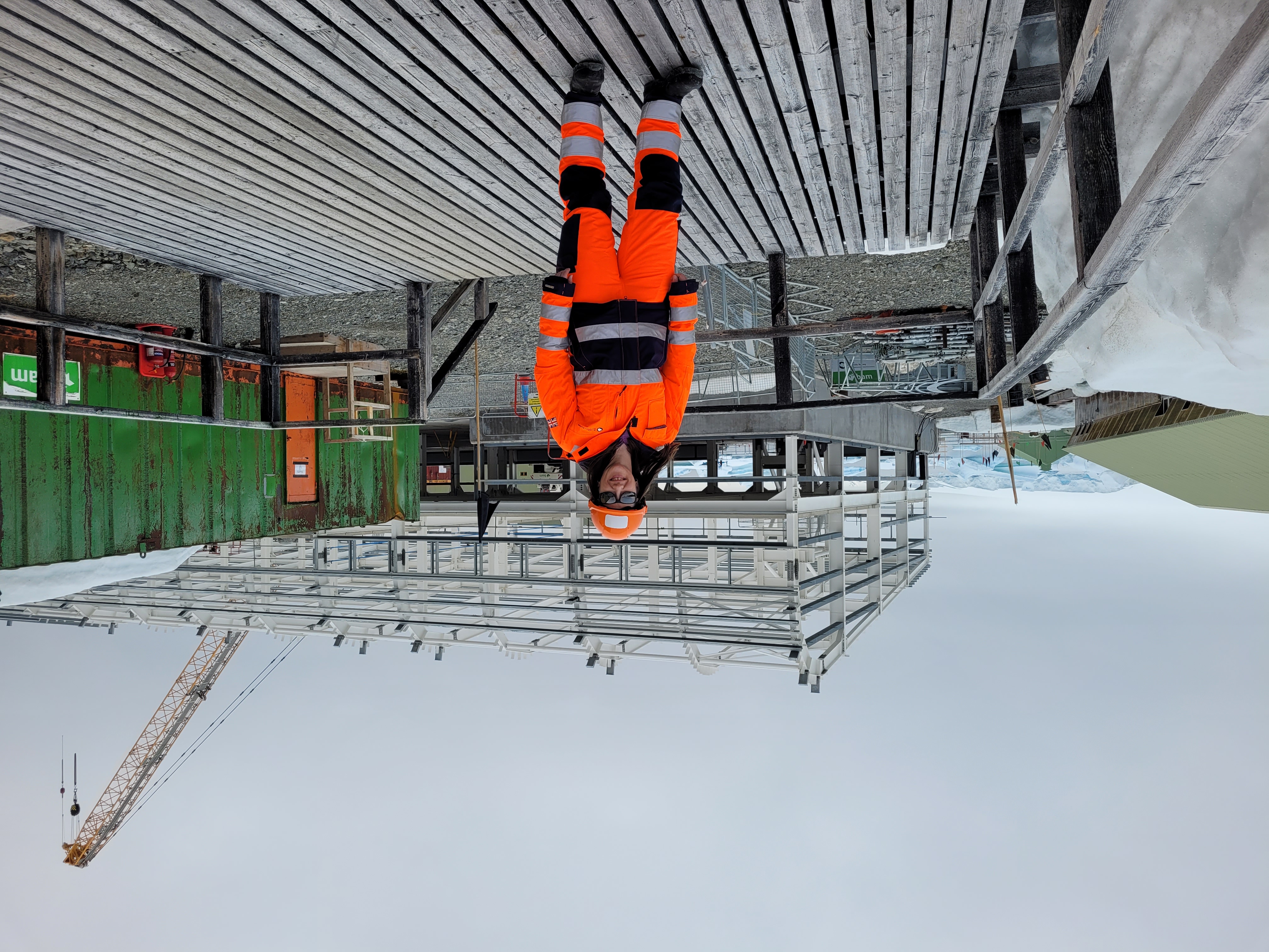 Elen Jones from BAS stands smiling on a wooden boardwalk in full orange hi-vis workwear, hard hat and sunglasses. Behind her is the part-built steel frame of the Discovery Building at Rothera, with a tall crawler crane, a green site container and stacked materials. Snow, gravel and distant sea ice frame the scene under a flat, overcast Antarctic sky.