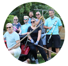 Here is a breakdown of the details in the image:  The People: The group is a mix of men and women of various ages. They are all smiling or looking pleasantly toward the camera. Many of them are wearing sunglasses.  The Clothing: The group is dressed in casual, practical outdoor clothing. A few individuals are wearing bright, light-blue t-shirts or polo shirts, while others are in black, grey, or navy shirts. Some members of the group are wearing protective work gloves.  Tools and Equipment:  A man on the right side of the frame is holding a large pair of loppers (tree trimmers).  A man in the back is holding up a large rake.  A woman in the foreground is holding a long-handled tool, possibly another rake or hoe.  A red riding lawn mower or utility vehicle is visible in the lower-left and centre, with a couple of group members positioned on or right next to it.  Setting: The background is filled with lush green trees and foliage, suggesting a park, large garden, or countryside setting on a bright, sunny day.