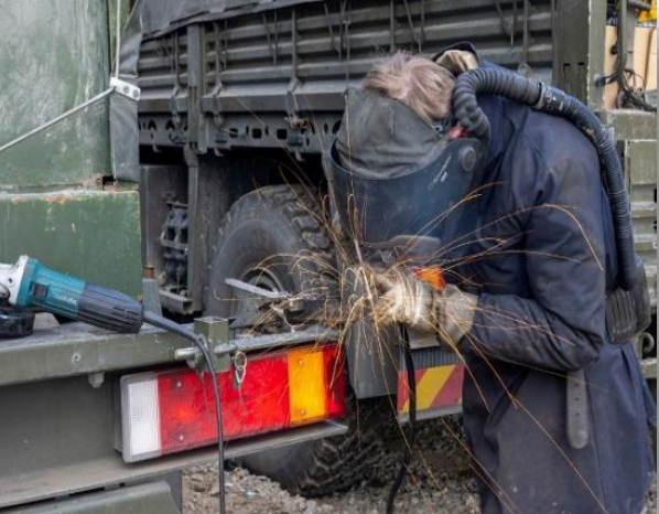 The image shows a person engaged in metalwork, likely grinding or cutting, on the back of a large military-style vehicle. The scene is filled with action and industrial detail.  Key Elements The Worker: A person wearing dark blue or black protective coveralls is leaning over a metal component. They are equipped with a welding helmet/face shield and heavy-duty gloves for protection against heat and sparks.  The Action: A shower of bright orange sparks is flying from the point of contact between a tool and the metal, indicating an abrasive process like grinding.  Equipment: * On the left, a teal and black angle grinder (likely a Makita brand based on the color and shape) rests on the edge of the vehicle bed.  The worker appears to be using a powered air-purifying respirator (PAPR) system, visible as a black corrugated hose connecting the back of their helmet to a battery pack on their waist.  The Vehicle: The work is being performed on the rear of a heavy-duty, olive-drab truck. Visible features include a large off-road tire, a metal tailgate, and a rectangular taillight assembly with red and amber sections.