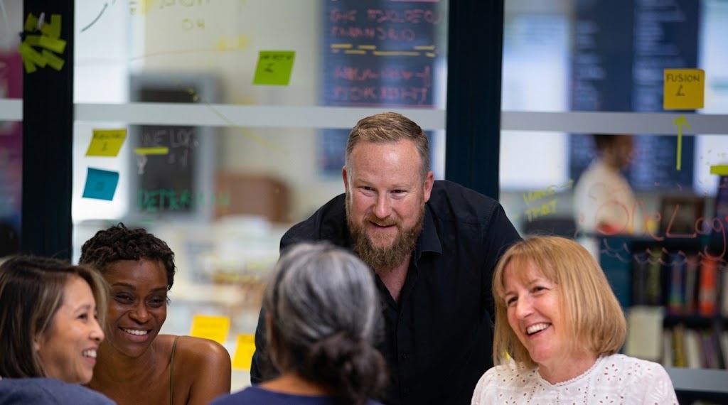 The image shows a collaborative workspace environment where a team is engaged in a discussion. Central Figures: At the centre of the frame, a man with fair hair and a full beard wearing a dark shirt leans forward with an engaged, smiling expression. In the foreground, four people sit around a table listening and participating in the conversation. People Present:On the left, a woman with short dark hair smiles toward the centre. Beside her, a Black woman with short dark hair smiles widely. In the centre foreground, the back of a woman's head is visible, showing greyish hair tied back. On the right, a woman with blonde hair and bangs, wearing a white textured top, laughs merrily while looking toward the group. Background & Setting: The group appears to be in an office or meeting area behind a glass partition. The glass is covered with colourful sticky notes (yellow, blue, and green) and handwritten notes or diagrams. The soft lighting creates a warm, professional, yet relaxed atmosphere.