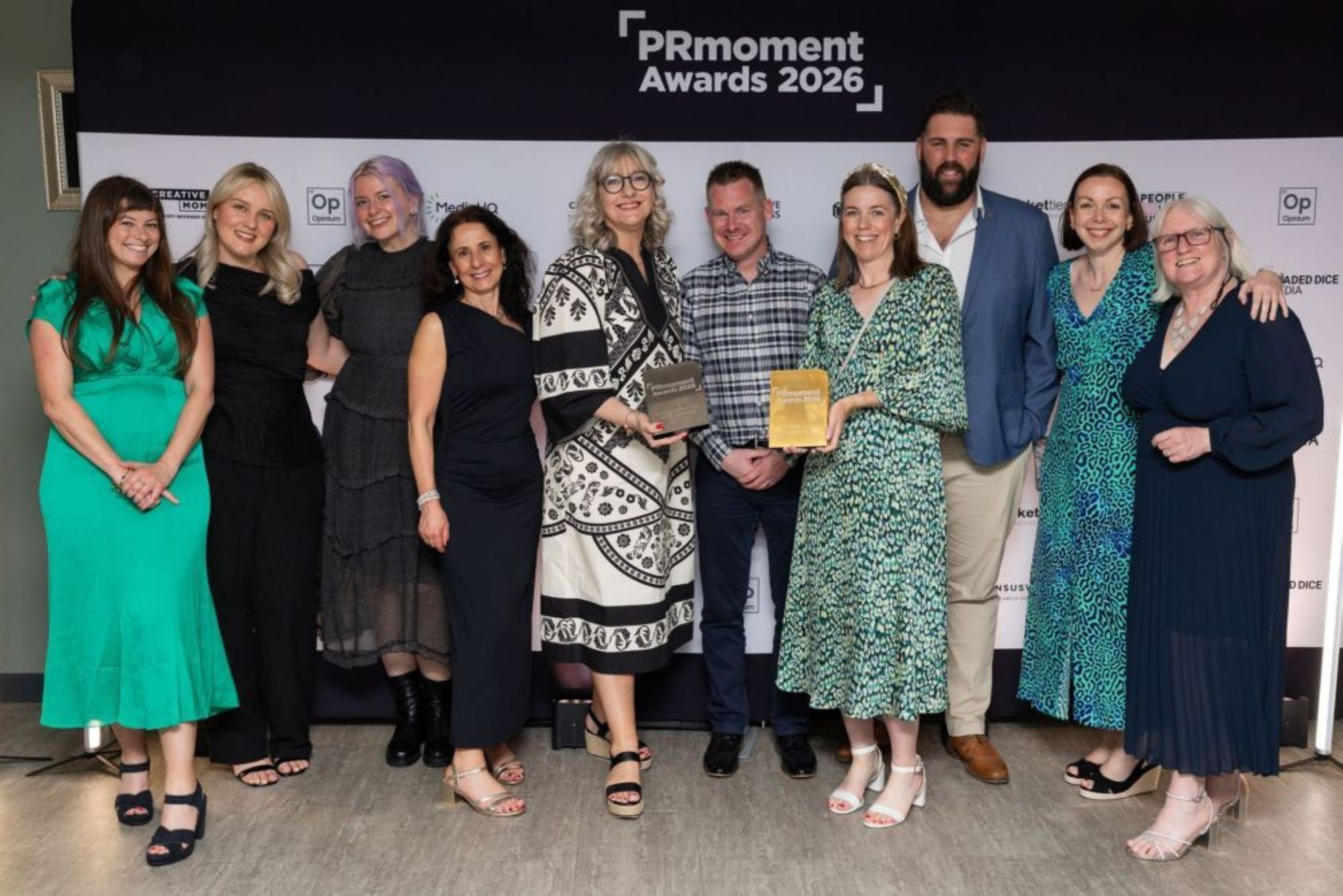 The Group: The group consists of seven women and three men, all dressed in smart-casual or formal attire. Their expressions are joyful and smiling, suggesting a celebratory atmosphere. The Awards: Two individuals in the center of the group are prominently holding awards. One is a dark, rectangular plaque held by a woman in a patterned black-and-white dress, and the other is a gold-colored rectangular award held by a woman in a green patterned dress. Setting: The photo is taken against a professional "step and repeat" banner. Bright studio lighting is visible on the floor at the edges of the frame, and the floor appears to be a light-colored wood or laminate. Fashion: The attire varies from vibrant colors, such as a bright green dress and a blue leopard-print dress, to classic black outfits and a blue blazer.