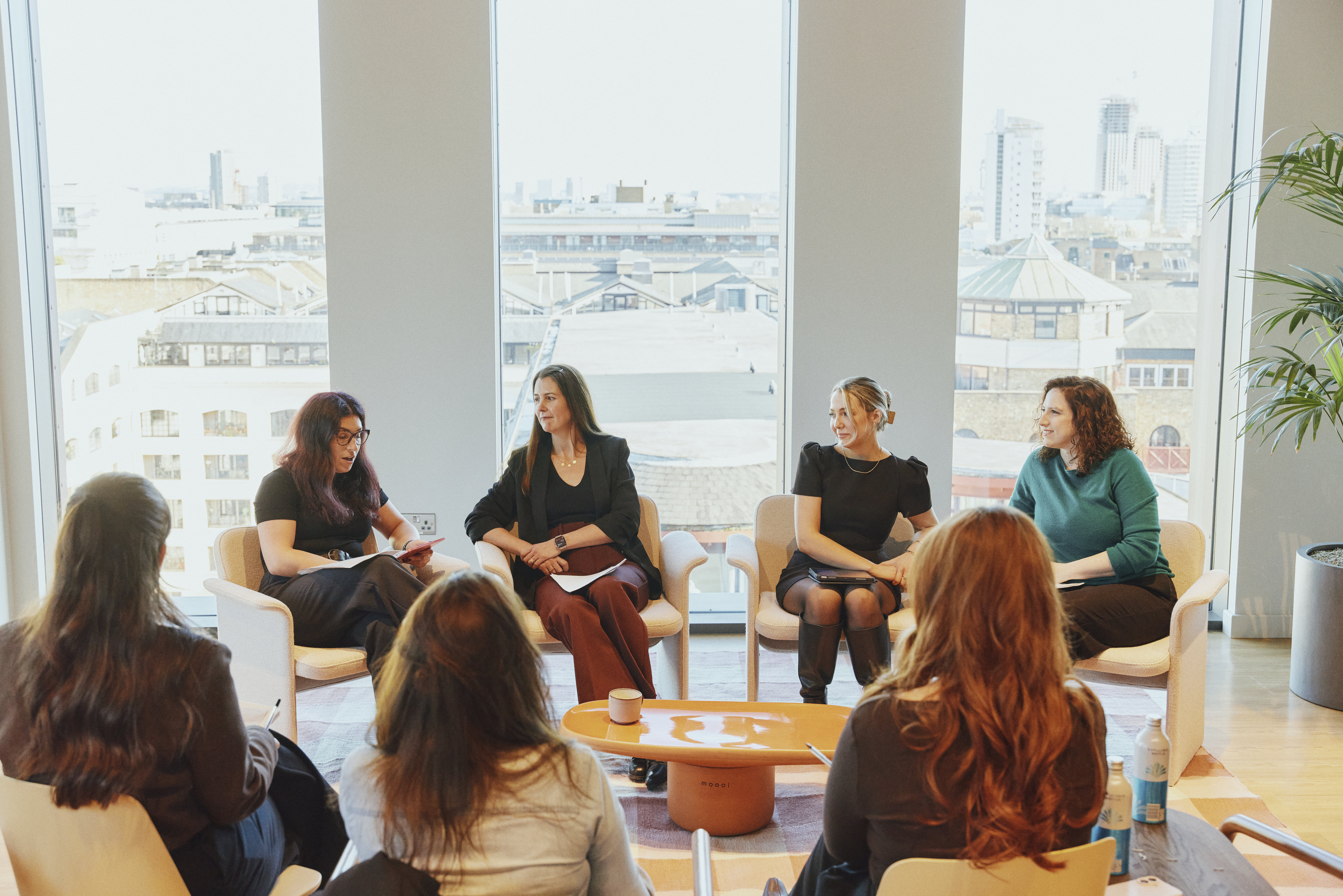 This image captures a professional panel discussion or workshop taking place in a modern, sunlit office space. A group of women are seated in a semi-circle, engaged in a collaborative and focussed conversation. Key Elements of the Scene The Participants: Four women are seated in plush, light-coloured armchairs facing the camera. On the far left, a woman in a black top is looking down at a red tablet or notebook. To her right, a woman in a black blazer and dark red trousers looks toward the centre of the group. Next is a woman in a black dress and boots, leaning forward slightly. On the far right, a woman in a teal jumper is smiling and looking toward the other speakers. The Foreground: Three other individuals are seated with their backs to the camera, suggesting an audience or additional participants in the circle. The Setting: The room features large, floor-to-ceiling windows that provide a panoramic view of a city skyline with various brick buildings and rooftops. Atmosphere and Decor: The lighting is bright and natural, creating a warm, professional "golden hour" glow. In the centre sits a low, orange oval coffee table with a single mug on it. A large potted palm plant is visible on the right, adding a touch of greenery to the minimalist interior. The overall layout and body language suggest an intimate, high-level meeting, panel talk, or corporate "fireside chat."