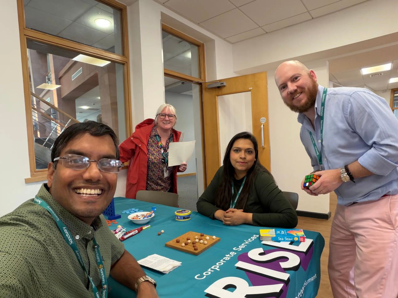 This image features four colleagues gathered around a display table indoors, in what looks like an office or corporate building setting. Key DetailsThe People:In the foreground on the left, a smiling man with glasses is taking a selfie-style shot wearing a patterned green shirt. Seated in the centre is a woman with long dark hair, wearing a dark green top. Standing behind her on the left is a woman in a red jacket holding a sheet of paper. On the right, a man with a beard and light blue shirt is leaning in towards the group, holding a Rubik's Cube. All four individuals are wearing green "Sanctuary" lanyards around their necks. The Display Table:Covered with a teal tablecloth featuring white branding text that reads "Corporate Services" and "RISE". On top of the table are various games and items, including a wooden Solitaire board, a Connect Four game set, small snacks/sweets, a bowl, and card games (including "Sea Snap!"). The Environment:Bright, modern indoor space with large interior glass windows, white walls, and a light-coloured wooden door frame
