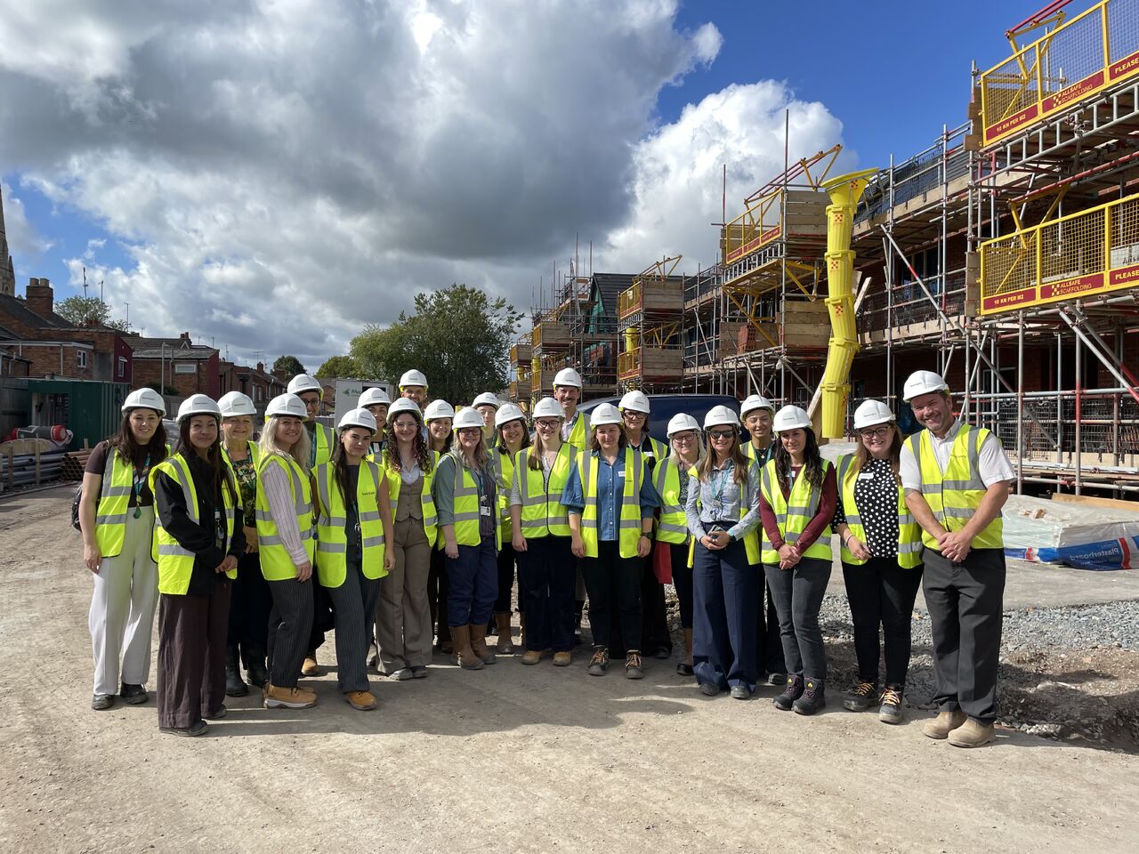 The image shows a large group posed together outdoors at a construction site. Key DetailsThe People: There are over 20 individuals, mostly women, lined up across an unpaved, gravel ground. Almost all of them are wearing white hard hats and high-visibility yellow safety vests over their smart-casual clothing. The Construction Site: Behind the group, a residential building under development is visible, completely clad in extensive yellow and metal scaffolding. A bright yellow rubbish chute extends vertically down the side of the scaffolding structure. Background & Surroundings: On the far left, existing residential houses and a church spire can be seen in the distance beyond the site boundary. Environment: The weather appears bright with a mix of blue sky and large white and dark clouds.
