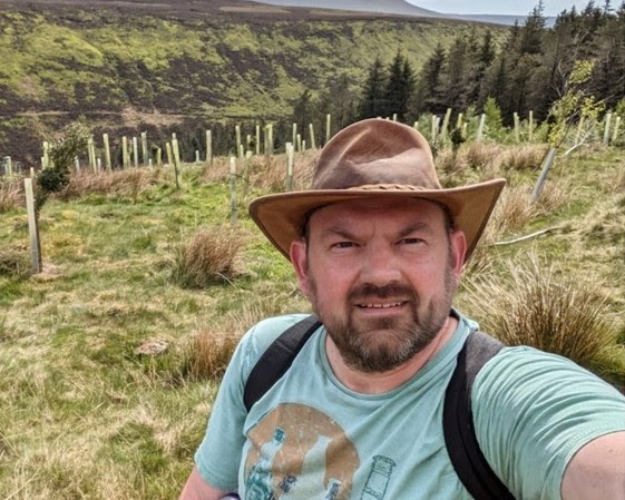 This image shows Tony Sutton in a rural, hilly landscape. He is a middle-aged man with a short beard and mustache, wearing a wide-brimmed brown hat, a light teal t-shirt with a graphic on it, and a black backpack. He is taking a selfie in what appears to be a valley or a rolling hillside. The background features grassy terrain, some pine trees, and rows of small saplings protected by light-colored tree guards. The weather looks bright and clear, capturing a great "outdoorsy" vibe that perfectly matches his Ben Nevis fundraising mission.