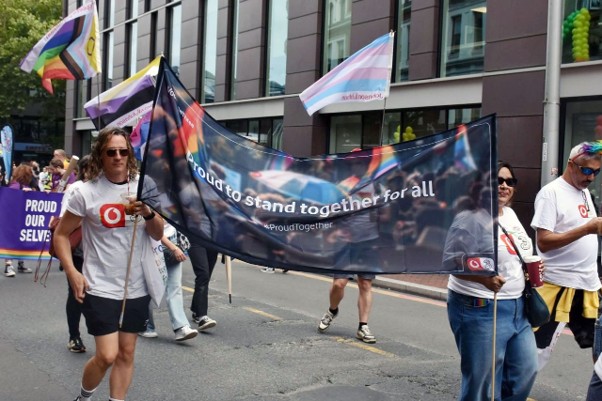 This is a photo of a Pride march taking place on a city street. A group of people are walking towards the camera carrying a large dark banner that reads "proud to stand together for all", with a smaller "#ProudTogether" hashtag beneath it. The banner has a faint photographic background and appears to be a VodafoneThree branded one. In the foreground on the left, a man in a white VodafoneThree t-shirt and shorts is holding one of the banner poles. He has light brown shoulder-length hair and is wearing sunglasses. Several other people in matching white t-shirts are carrying the rest of the banner and walking alongside it, including a person on the right using a long white cane. The background is full of Pride imagery: a Progress Pride flag and a Transgender Pride flag (light blue, pink and white) are flying on the left, and more rainbow and trans flags are visible on the right. To the left edge there's another purple banner, partly cut off, with white text that includes the words "PROUD" and "OUR" and "SELVES", suggesting something like "proud to be our [whole] selves". The setting is a paved road lined with modern buildings, with what looks like a balloon arch in the top right corner. The lighting is bright and overcast, and the overall mood is celebratory.