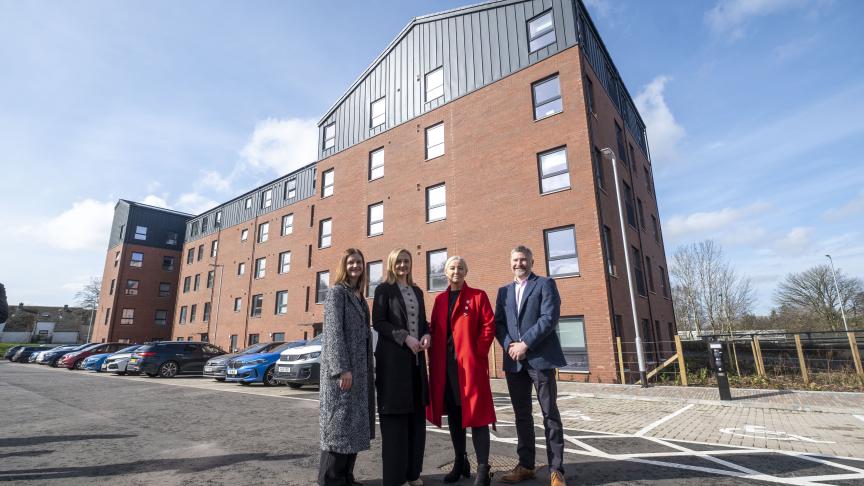 This image shows a wide-angle, outdoor shot of four people standing in front of a modern, multi-story apartment building. The Setting The building is a large, red-brick structure with five floors. It has a distinctive architectural style: The top section and some side panels are clad in dark grey or black corrugated metal. The windows are uniform, framed in dark grey, and reflect the bright daylight. The building sits behind a paved parking area with several cars (blue, white, and dark grey) parked in a row to the left. The ground in the foreground is asphalt, marked with white lines for parking spaces, including a designated disabled parking bay. The People Four people are standing together in the center-right of the frame, positioned as if posing for a professional or promotional photograph. From left to right: A woman wearing a long, grey patterned wool coat and black trousers. A woman in a long black coat worn over a beige top and black trousers. A woman wearing a vibrant, knee-length red coat over dark clothing, which stands out as the brightest element in the image. A man in a navy blue blazer, a light-colored collared shirt, and dark trousers.