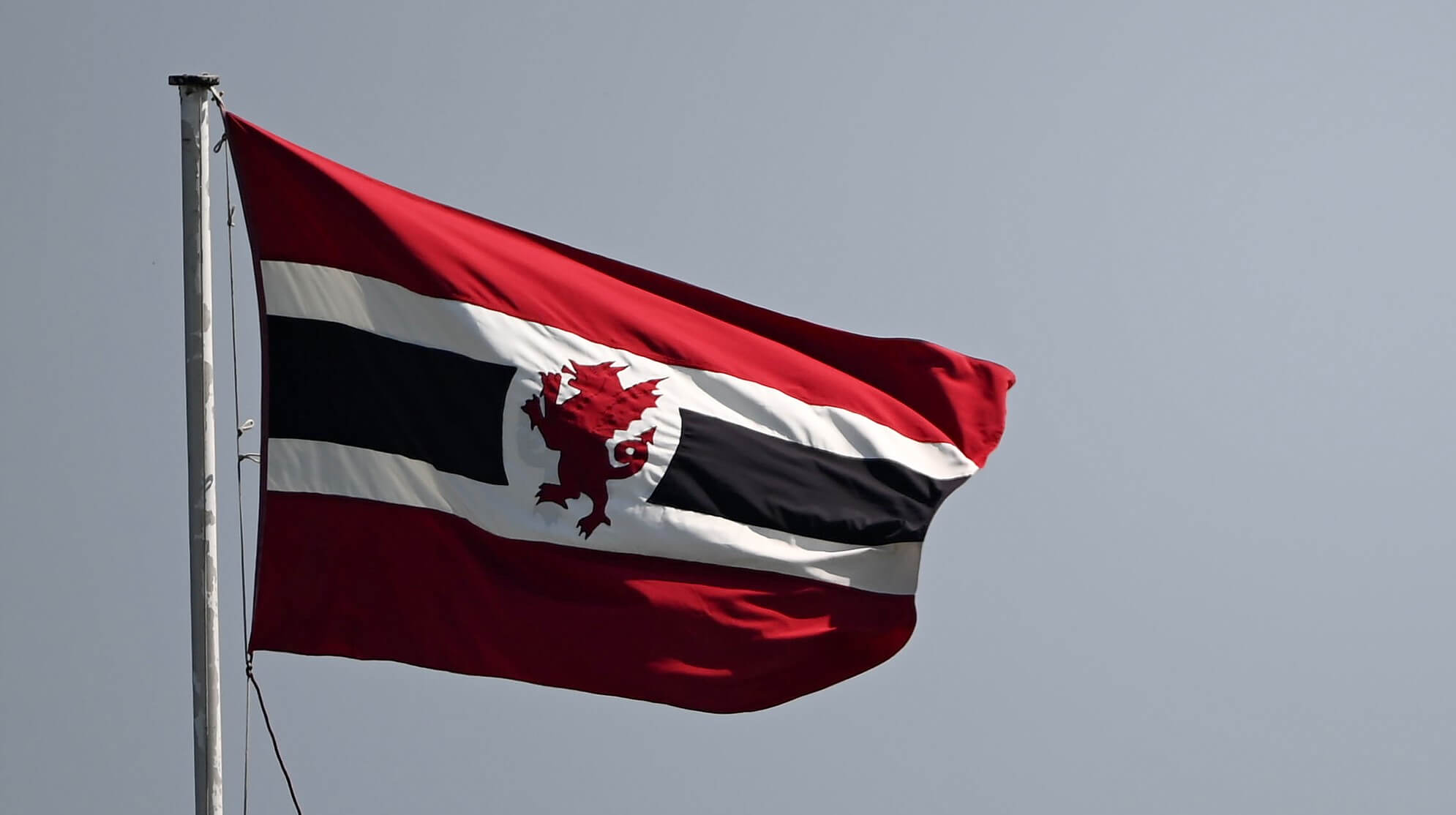The image shows a flag flying against a plain, overcast, light-grey sky. Flag Design: It features a deep red background with a prominent horizontal central band running across the middle. This central band consists of a thick black horizontal stripe bordered by narrow white fimbriation top and bottom. Emblem: In the centre of the flag, breaking the horizontal black and white band, is a silhouette of a red dragon (a griffin or wyvern style heraldic beast) positioned on a white vertical background block/circle cutout. Details: The flag is attached to a light grey metal flagpole on the left side and is waving gently in the breeze towards the right, displaying natural shadows and creases across the fabric.