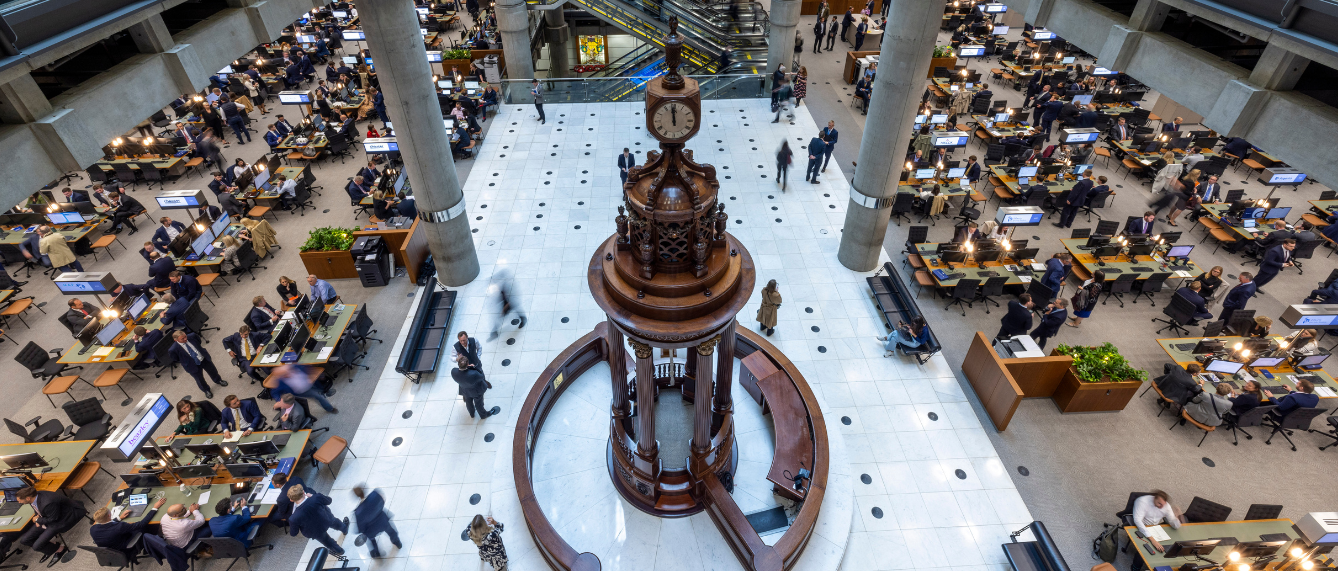 This image captures the iconic Underwriting Room at Lloyd’s of London, characterized by its "inside-out" high-tech architecture and exposed concrete pillars. At the heart of the bustling marketplace stands the ornate wooden Lutine Bell rostrum, a historic centerpiece surrounded by modern underwriting desks where professionals gather to trade risk. The scene illustrates a blend of tradition and modern technology, perfectly reflecting the "professional, collaborative, and friendly" culture highlighted in the 2025 survey.
