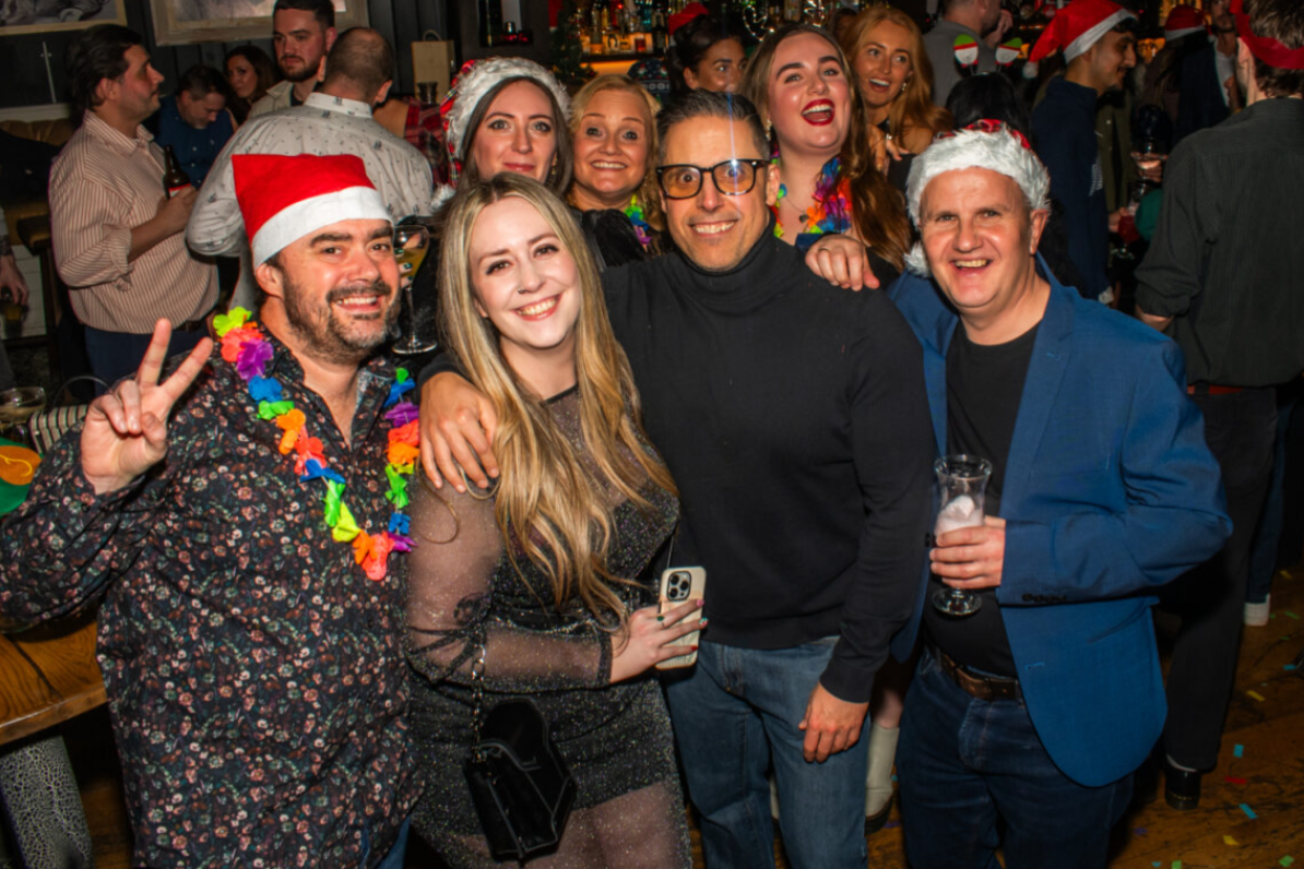 A crowded, brightly lit indoor venue, likely a bar or pub during a festive celebration, is shown in a medium full shot. Four individuals stand close together in the foreground, posing for a photo, while several others fill the background.  The individual on the far left, a person with dark hair, a beard, and a moustache, wears a red and white Santa hat, a dark, floral-patterned button-down shirt, and a colourful, rainbow-coloured flower lei around their neck. Smiling warmly, they raise their right hand in a peace sign.  To their right, another person with long, blonde hair smiles brightly. They wear a dark, shimmering, semi-sheer long-sleeved dress and hold a smartphone in their left hand while a small black purse hangs from their shoulder.  Beside them, a person with short, dark hair wears rectangular glasses with dark frames and a black turtleneck sweater with blue jeans. They smile broadly, their arm resting on the blonde person&rsquo;s shoulder.  On the far right, a person with short, light-brown hair wears a red and white Santa hat, a dark t-shirt under a blue blazer, and blue jeans. They smile, holding a glass of a clear beverage with foam in their left hand.  The background is filled with several other people, some wearing festive attire like Santa hats or novelty headbands, all appearing to be enjoying a lively, social gathering. The lighting is warm and direct, highlighting the cheerful atmosphere of the event.