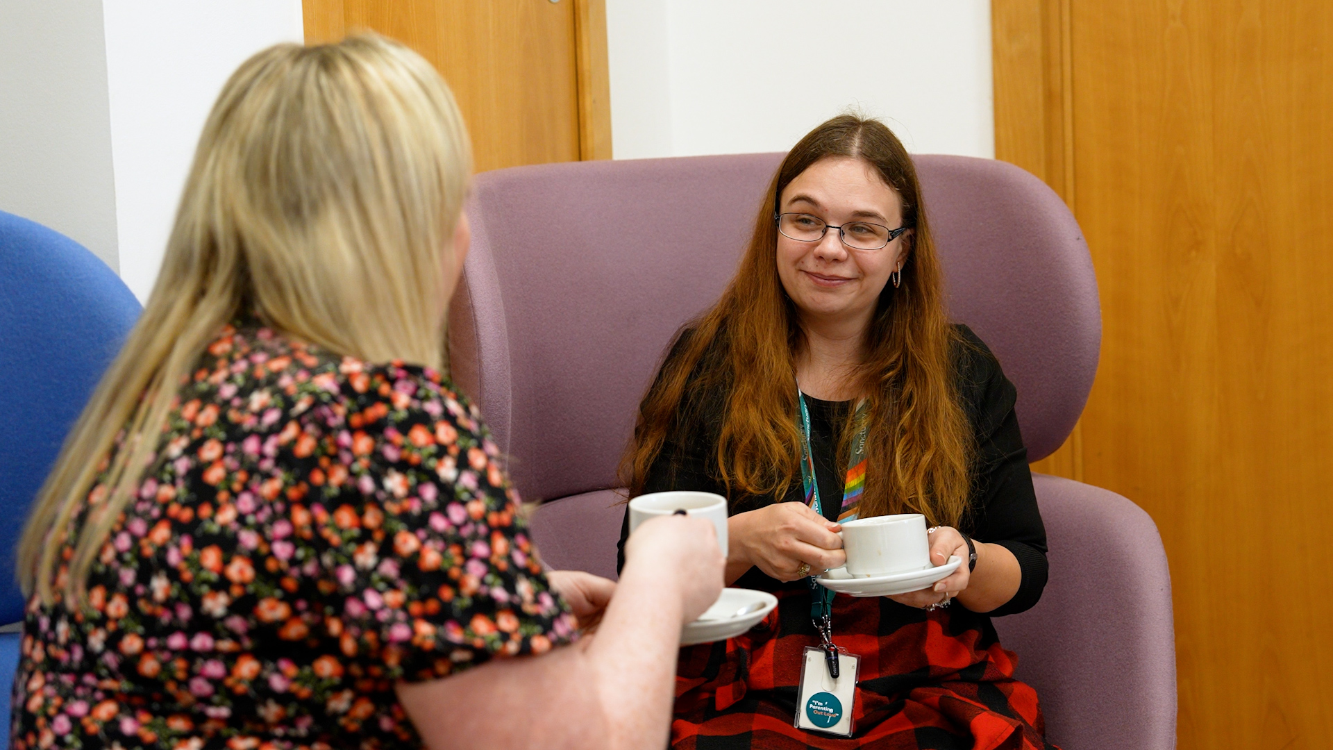 Gemini said The image shows two people engaged in a friendly conversation in a professional yet comfortable indoor setting. One person, seen from the back, has blonde hair and is wearing a colorful floral-patterned top. The other person is seated in a large, purple high-backed armchair, wearing glasses and a black top with a red-and-black checkered skirt. They are smiling warmly while holding a white teacup and saucer. The person in the chair also wears an ID badge on a lanyard, suggesting an office or community service environment. Both individuals appear to be enjoying a coffee or tea break together.