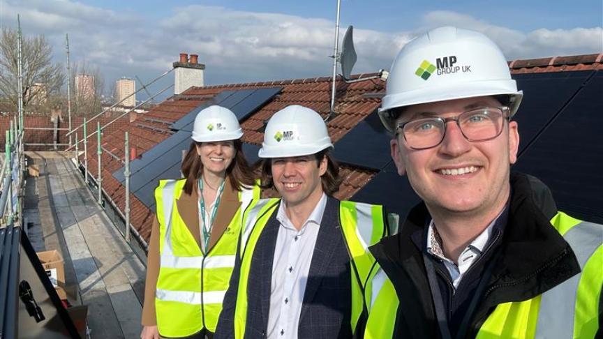 This image shows three people posing for a selfie on a rooftop construction site, likely documenting the progress of a renewable energy project. The People Three individuals are featured in a close-up, smiling toward the camera. They are all wearing safety gear, including white hard hats and high-visibility yellow vests. The hard hats display a logo that reads "MP GROUP UK." The person in the foreground (on the right) is wearing glasses and holding the camera for the selfie. The two people in the background are standing on a wooden scaffolding platform that runs alongside the roof. The Setting The Roof: The group is standing next to a sloped roof with traditional reddish-brown clay tiles. Large, black solar panels have been installed across the tiles. Infrastructure: Scaffolding with green metal poles and wooden planks is visible on the left side, indicating active construction or installation work. A satellite dish and a brick chimney are also visible on the roofline. The Background: In the distance, you can see a city skyline with several high-rise residential buildings under a bright, slightly hazy blue sky with light clouds.