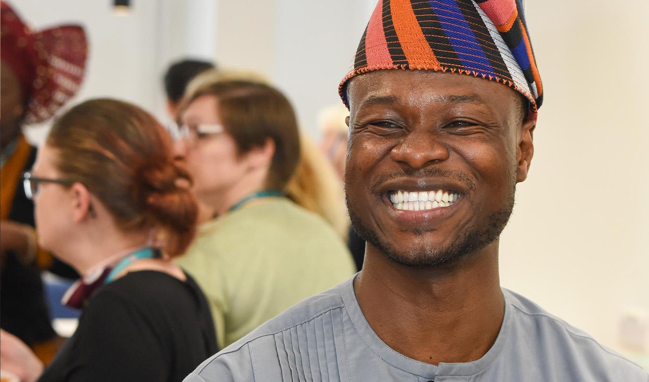 The image shows a man smiling broadly at the camera. He is wearing a colourful traditional West African cap, commonly known as a "fila" or Yoruba cap, featuring stripes of orange, blue, black and white. He is also wearing a grey traditional top that appears to be a dashiki or similar West African garment with vertical pleat detailing on the chest. The setting looks like an indoor social gathering or event, with several people visible but out of focus in the background.