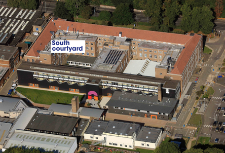 Key Visual Elements Main Building Structure: A large, U-shaped perimeter building featuring brick walls and red tiled pitched roofs, forming an inner quadrangle. Modern Extension: Running along the front of the complex is a long, modern building with dark (black) exterior cladding, horizontal ribbon glazing, and a bright pink archway entrance at the centre. Courtyard Area: A sloped green lawn area sits directly in front of the modern black wing, adjoining paved access pathways and additional lower-rise facilities. Roof Details: Various sections display flat roofs fitted with solar panels, skylights, and mechanical plant equipment. Surrounding Campus: The complex is surrounded by internal access roads, parking bays, pedestrian footpaths, and surrounding green trees along the top edge of the frame. Text Overlay: A prominent white label with blue text in the upper-left quadrant marks the area as: "South courtyard"