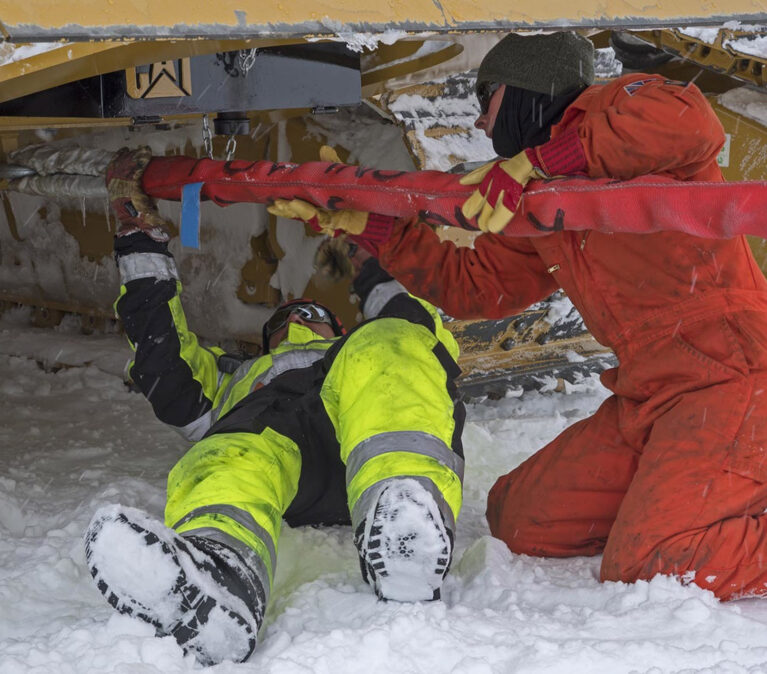 Two BAS vehicle mechanics work on a large yellow tracked vehicle in the snow. One, in yellow and black hi-vis waterproofs, lies flat on their back on the ground, sliding underneath the machine to reach it from below. The other, kneeling in an orange boiler suit with heavy gloves and a face covering, feeds a red strap or sling across to them. Both are heavily wrapped against the cold, with snow falling and settling on their clothing. The underside and track of the vehicle fill the top of the frame.