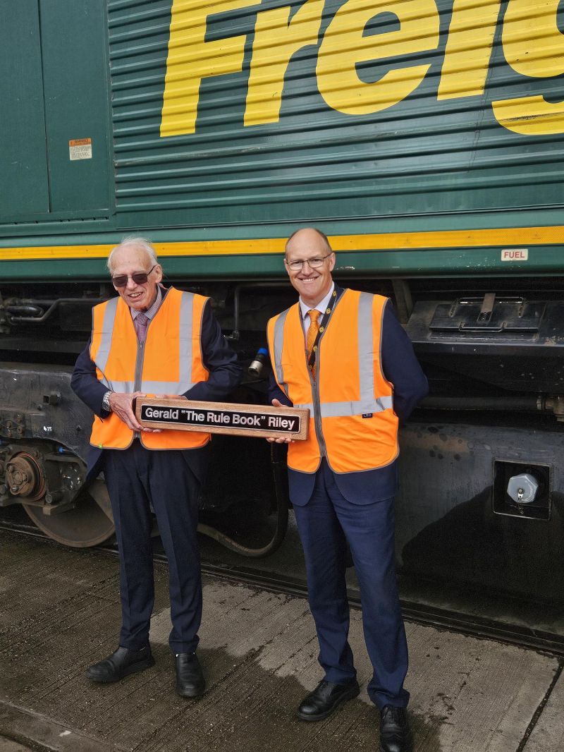 Gerals standing in front of a freight train with a collegue and holding a sign that reads 'Gerals 'Rule Book' Riley'