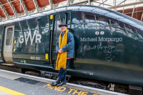Michael Morpurgo, a white middle aged man wearing denim jeans and jacket with a yellow scarf