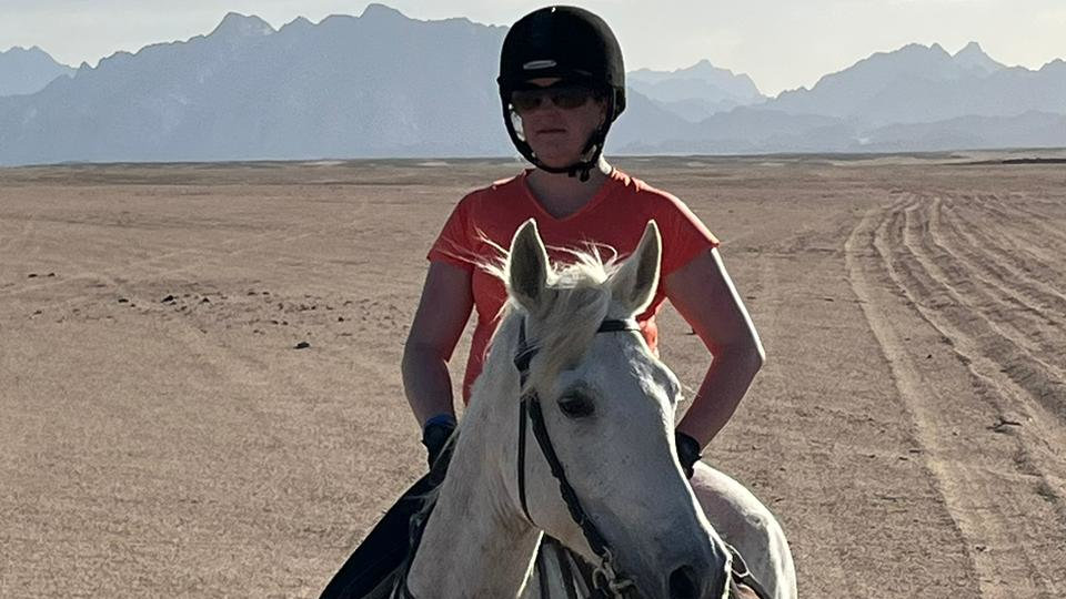 Ruth riding a white horse with a black riding hat and orange t-shirt