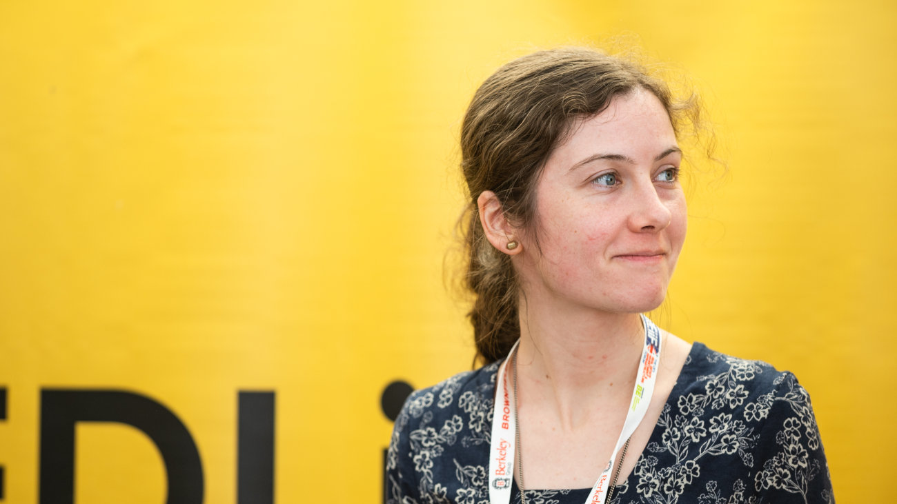 Rachel has brown hair, tied in a pony tail and is in front of a bright yellow background