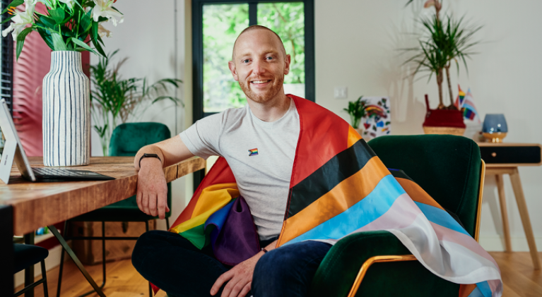 White smiling male sitting down with a pride flag around their shoulders