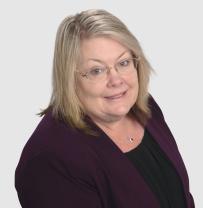 Head and shoulder hot of a white female looking up to the camera smiling. She us wearing a black top with a purple jacket. She has shoulder length blonde hair and glasses