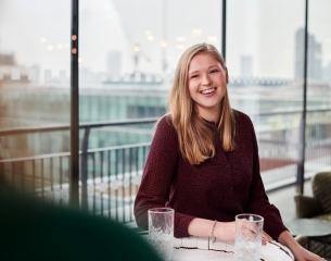 White female with long blonde hair sat at a table in front of a window smiling