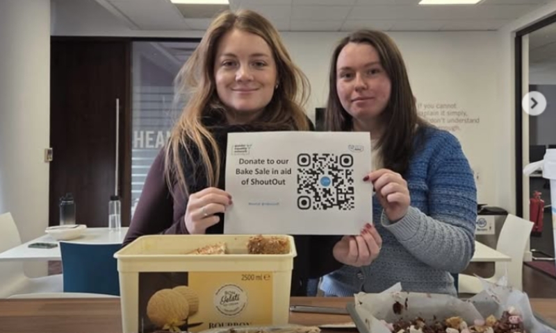 two white women hold a sign in front of bake sale