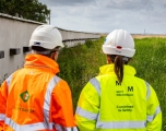 Two peoplestanding facing a field,one male one female, wearing branded high-vis jackets
