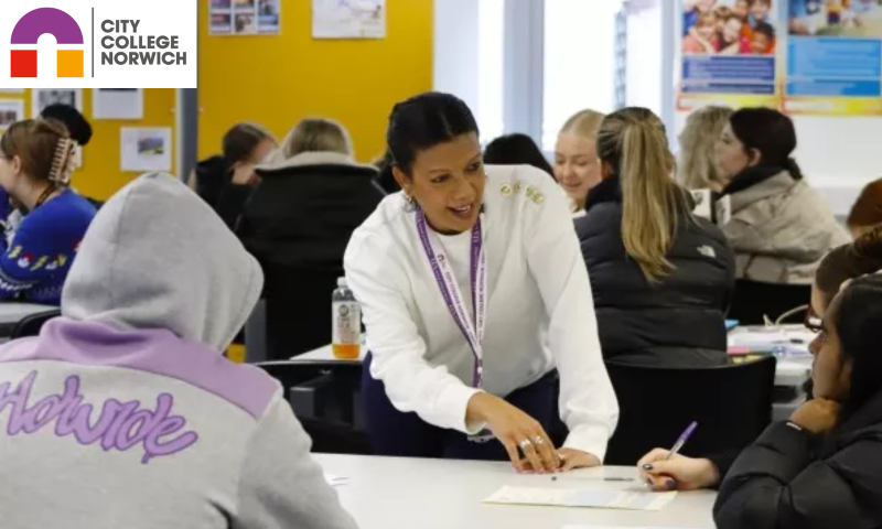 A female educator wearing a white sweater and a purple lanyard leans over a table to assist a group of students in a bright, modern classroom.