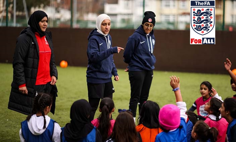 The scene takes place outdoors on a green artificial turf football pitch. In the center, three female coaches are leading a session. They are standing and facing a group of young girls who are sitting on the grass in a semi-circle, listening attentively.