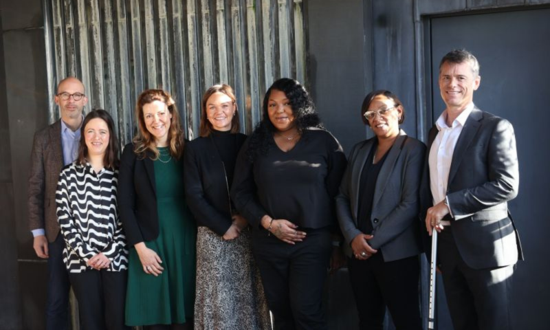A group of seven people, dressed in professional business attire, are standing together and smiling for a portrait in front of a textured gray wall. The group consists of five women and two men, including a man on the far right who is holding a white cane.