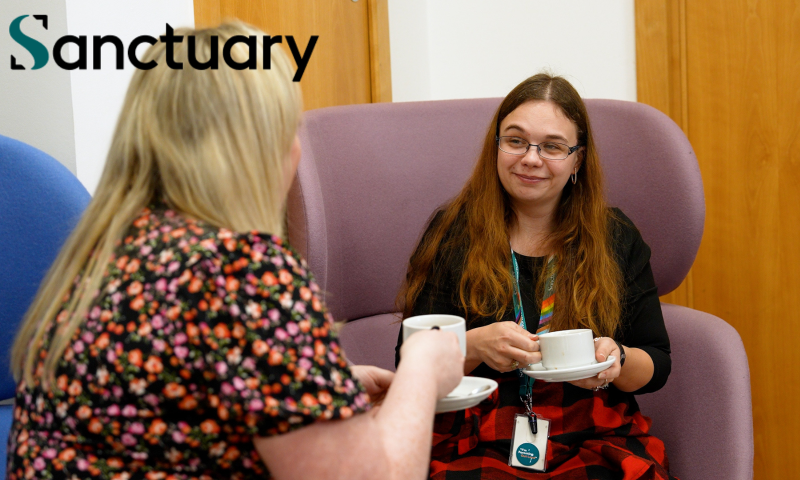 Gemini said The image shows two women sitting in comfortable chairs, having a conversation over tea or coffee. One woman is facing the camera with a slight smile, wearing glasses and a red and black patterned skirt, while the other is seen from the back, wearing a floral-patterned top.  In the top left corner, the word "Sanctuary" is visible in a bold, dark font. The setting appears to be a warm, indoor environment, likely a common room or a lounge area, emphasizing a relaxed and supportive atmosphere.
