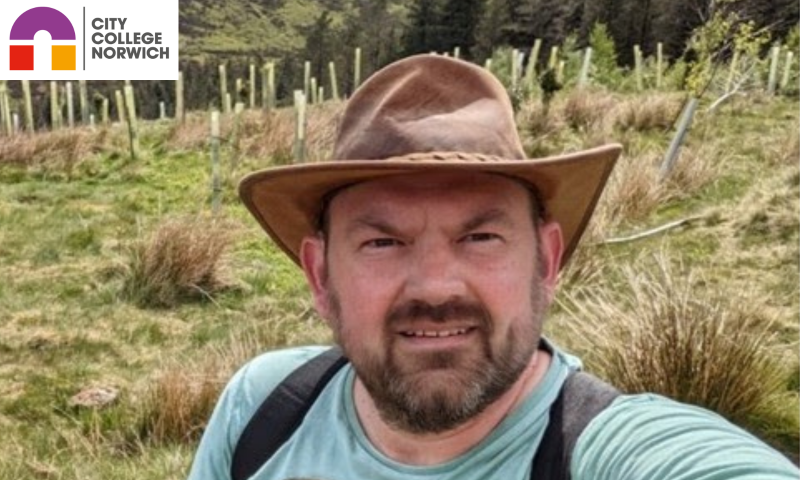 This image shows Tony Sutton in a rural, hilly landscape. He is a middle-aged man with a short beard and mustache, wearing a wide-brimmed brown hat, a light teal t-shirt with a graphic on it, and a black backpack.  He is taking a selfie in what appears to be a valley or a rolling hillside. The background features grassy terrain, some pine trees, and rows of small saplings protected by light-colored tree guards. The weather looks bright and clear, capturing a great "outdoorsy" vibe that perfectly matches his Ben Nevis fundraising mission.
