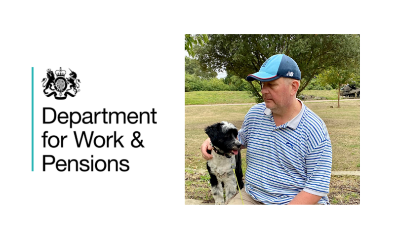 David is shown outdoors in a park-like setting with green trees and grass in the background. He is wearing a blue and white striped polo shirt and a blue baseball cap, and he has his arm around a small, fluffy black-and-white dog